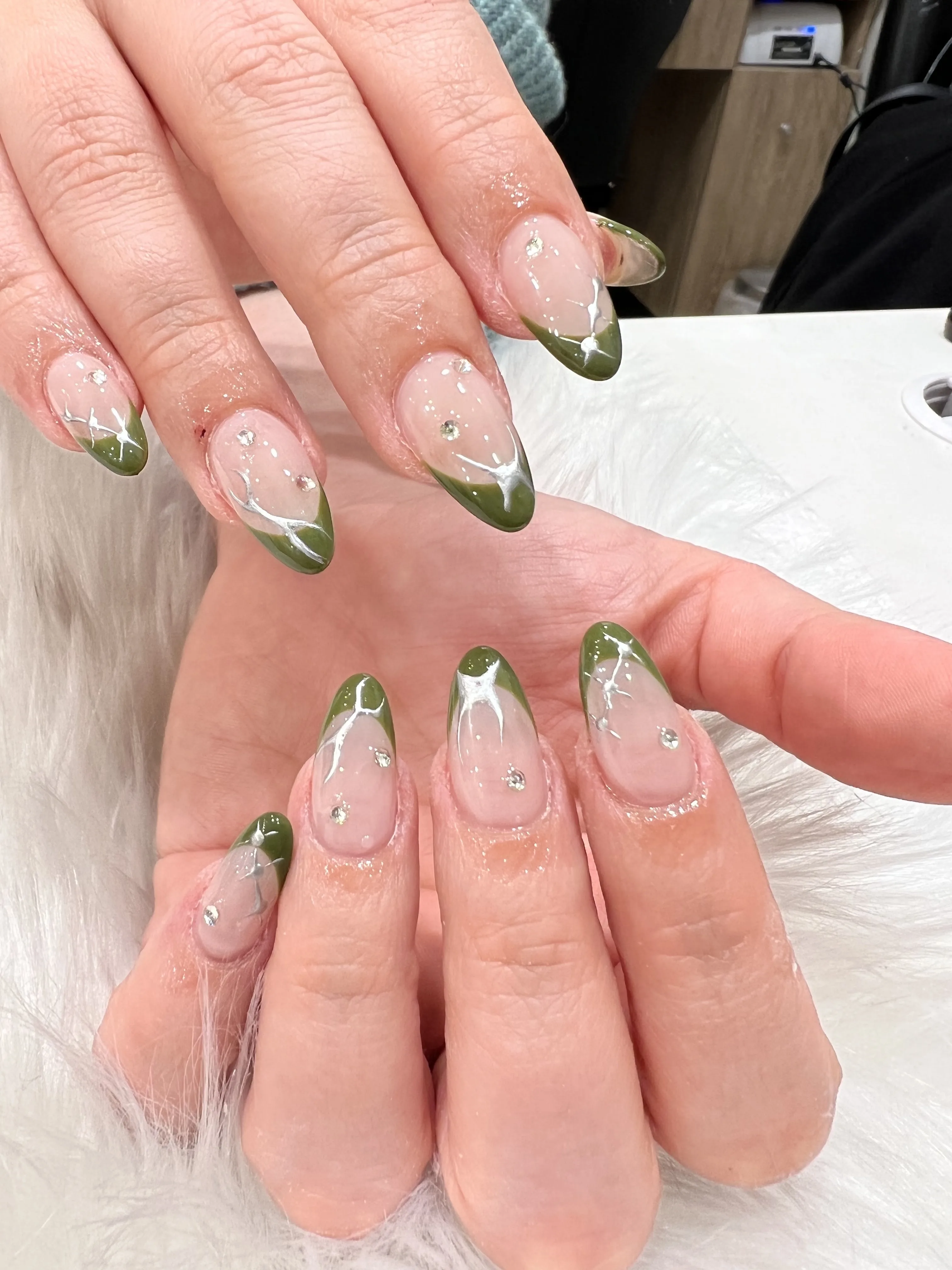 Olive-green French almond nails with silver chrome accents and gems at a nail desk
