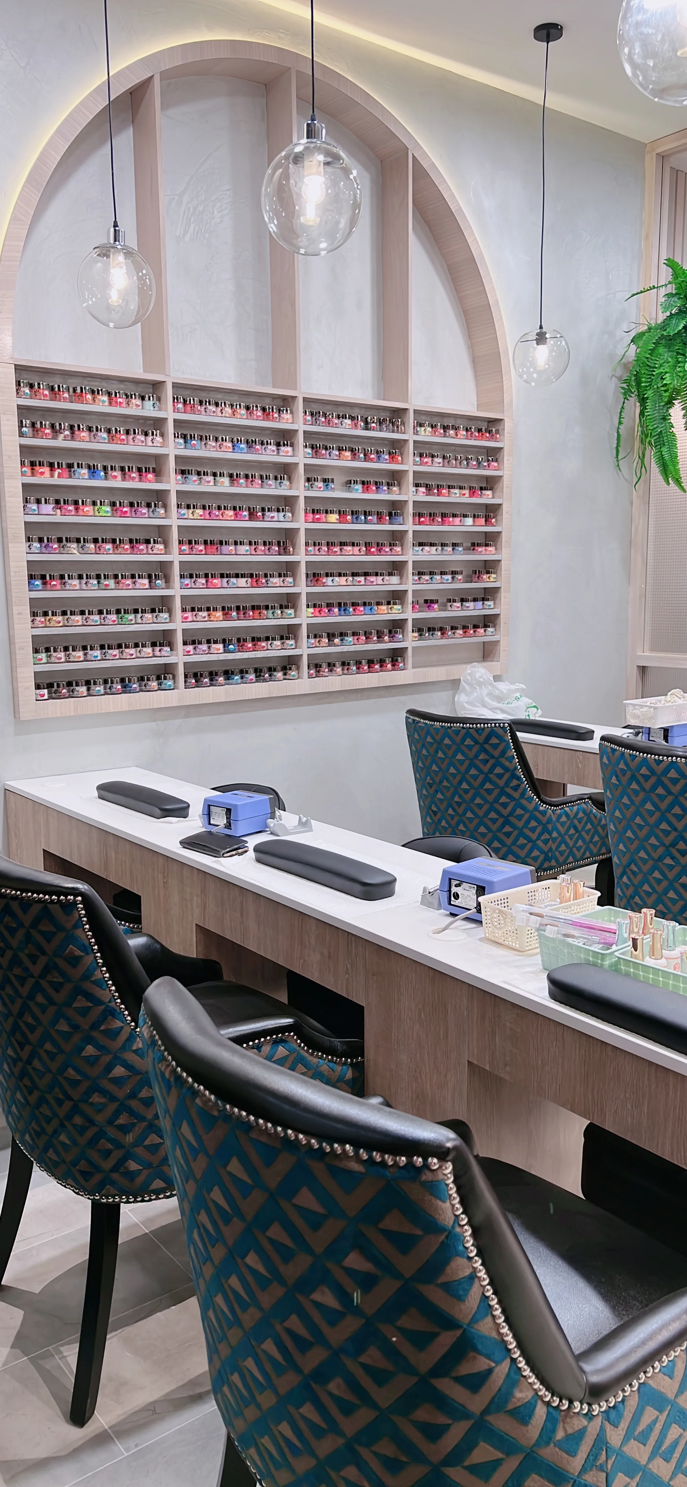 White-marble manicure desks with teal diamond-patterned chairs and a full wall of nail-polish colour under an arch with hanging ferns